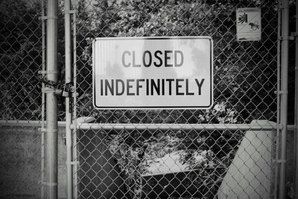 A black-and-white photo of a chain-link fence with “Closed Indefinitely” sign and padlock.