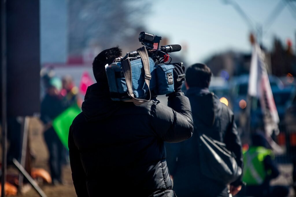 Man carrying a professional camera with people holding signs in the background.