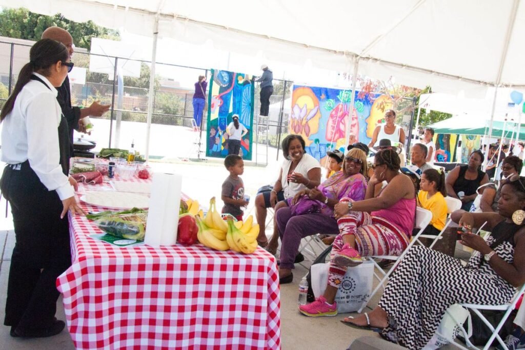A group of mostly Black women listen to a food lecture under a tent. In front, two people stand at a table of food items.