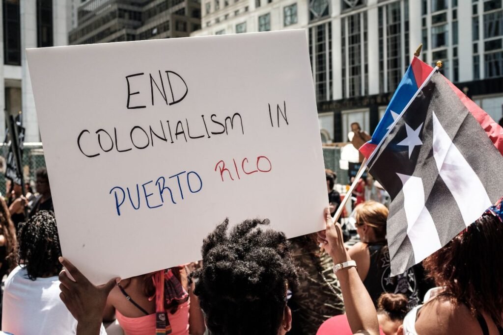 A person holds a sign that says “End colonialism in Puerto Rico. National and resistance Puerto Rican flags are visible.