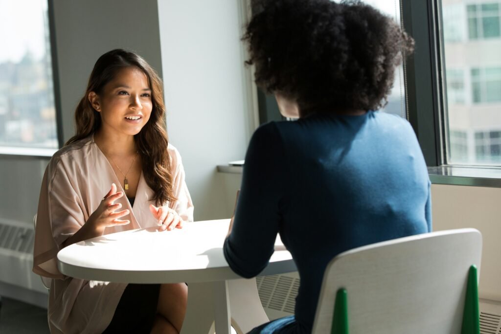 A woman sits across from another woman at a small table in conversation.