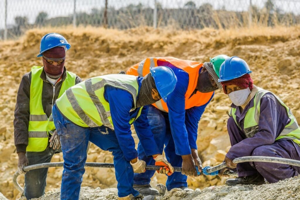 Four workers, all wearing masks, hard hats, and protective vests, hold hoses and work on a construction project together.