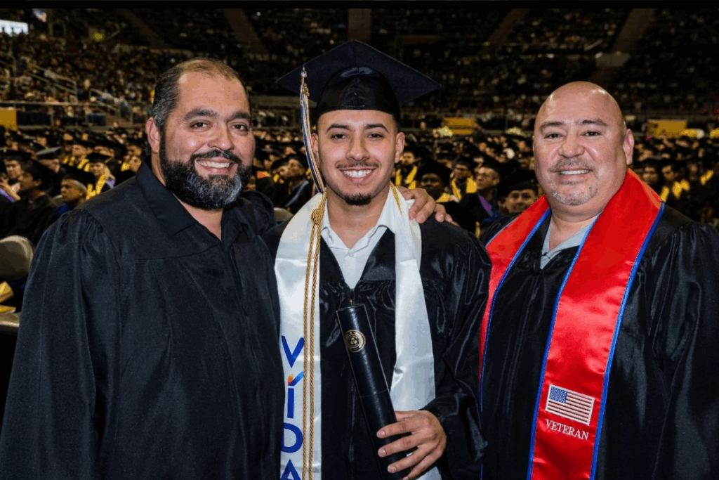 A picture of a graduate wearing a VIDA stole around his neck and surrounded by two other people.