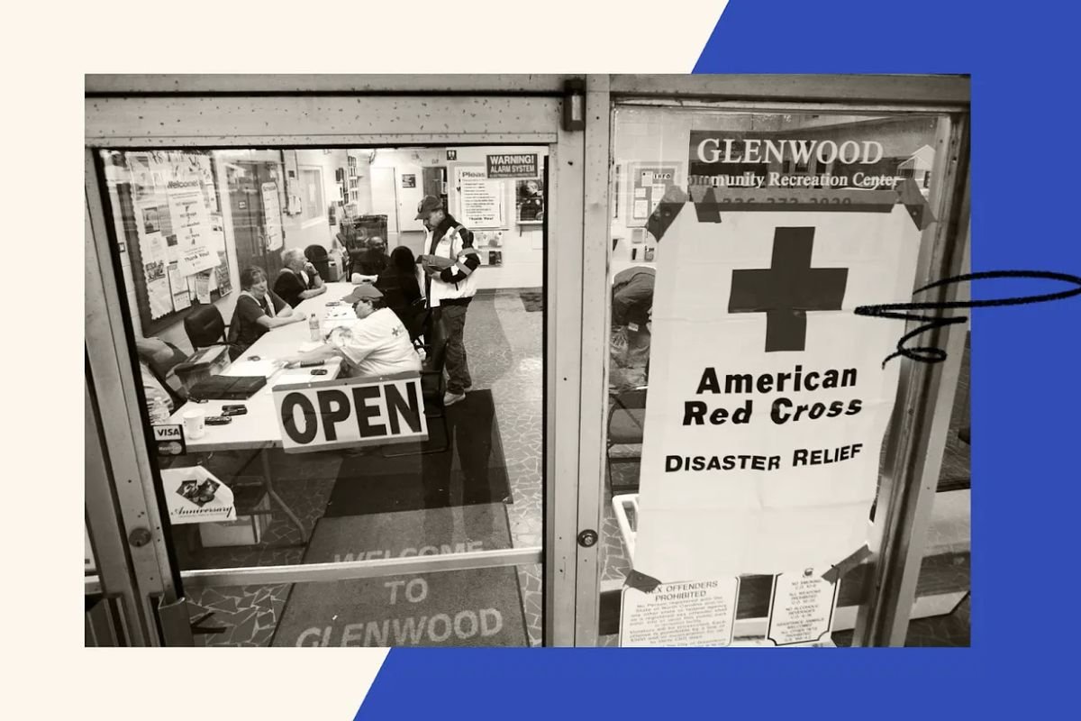 A black and white photo looking through a glass door into a room where people are sitting at a table. Signs on the door read "Glenwood Community Recreation Center," "American Red Cross Disaster Relief," and "Open".