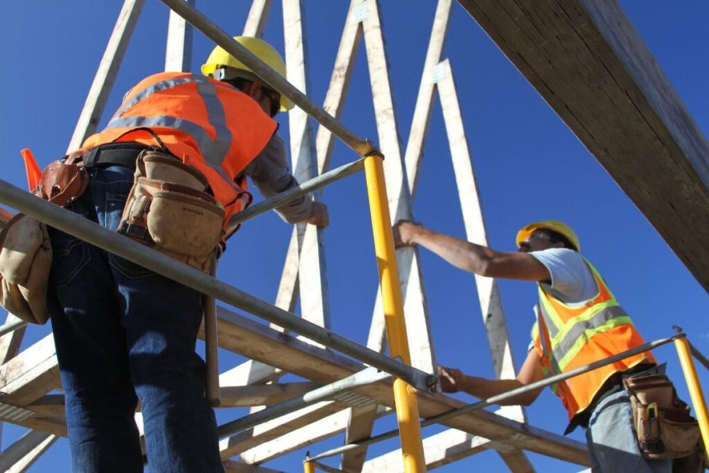Two construction workers, both wearing blue jeans, orange vests, and yellow hard hats, install part of a building frame.