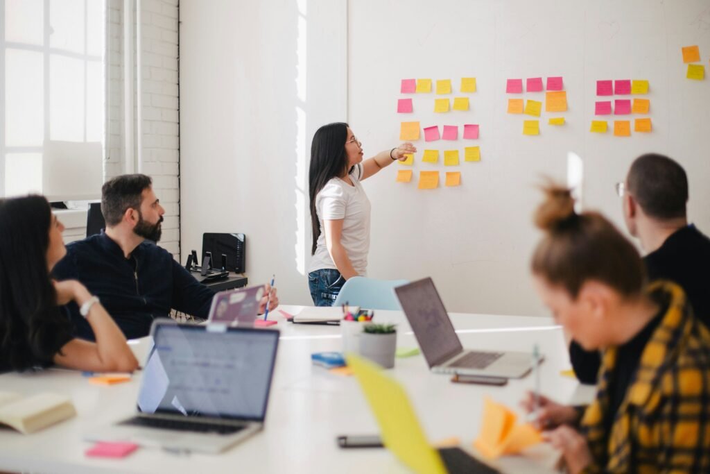 Woman in front of the group, reviewing sticky notes posted on a dry-erase board.