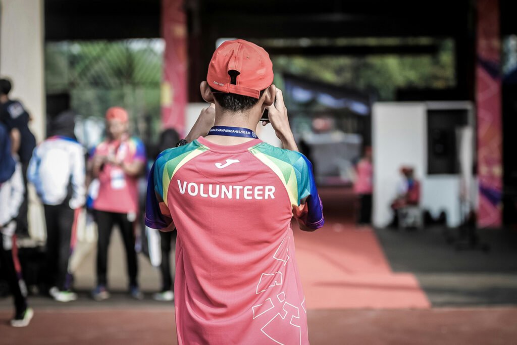 A young man in Indonesia wearing a shirt that reads, “Volunteer” and taking a picture of a building. He is photographed from behind.