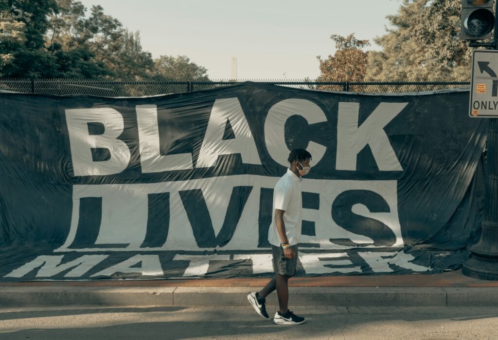A young Black man walks down a road with a large “Black Lives Matter” tarp hoisted behind him.