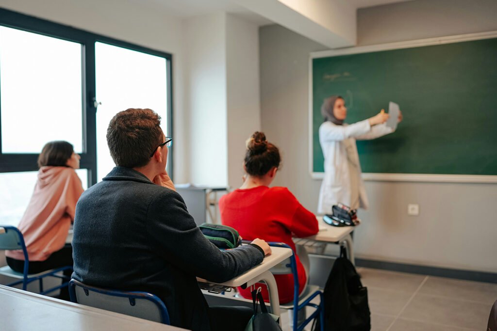 A hijabi woman professor standing in front of a class of college students and giving a lecture.