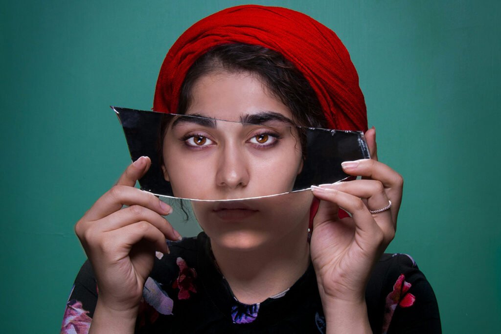 A south asaian woman with a red headwrap holding up a mirror shard that shows another woman’s face aligned with her own.