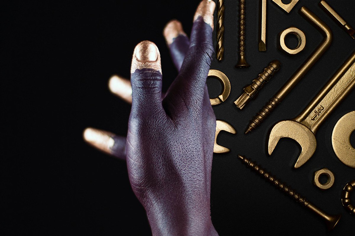 A Black hand with golden finger tips reaches upward, behind the hand there is an arrangement of golden tools.