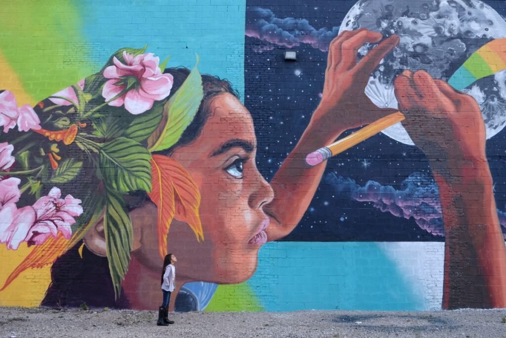 Mural of a child with flowers in their hair, drawing on the moon with a pencil, watched by a real child standing below, blending nature and imagination.