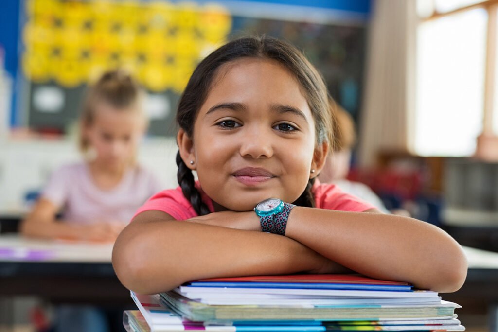 A young Latina girl with two braids rests her head on a stack of books in a classroom and smiles at the camera.