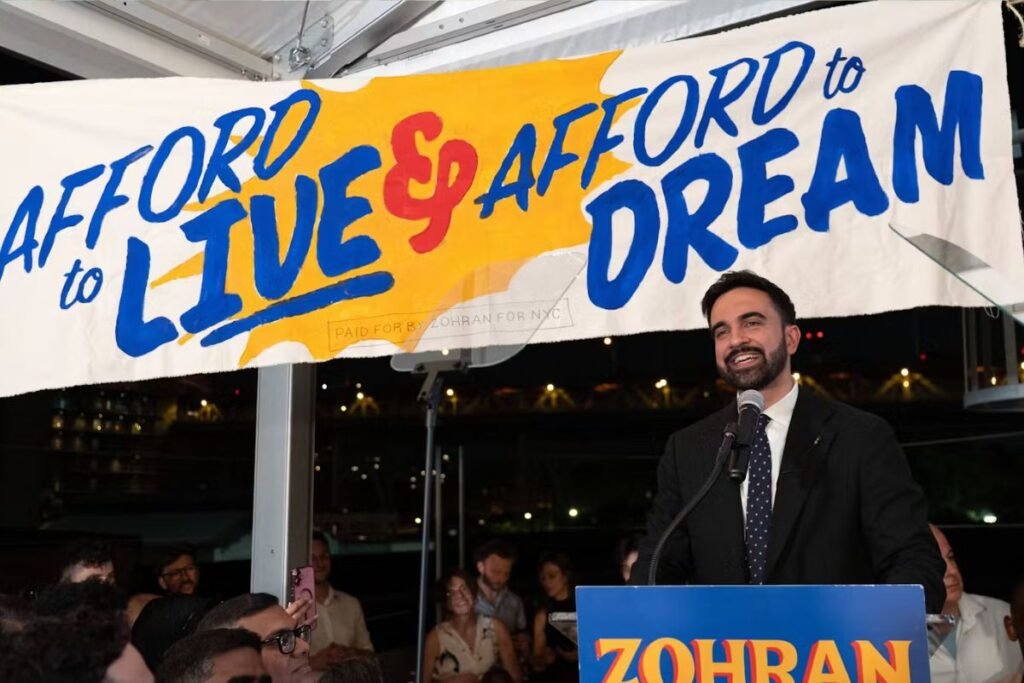 Zohran Mamdani speaks at a rally. Behind and above him is a large banner that says: “Afford to live and afford to dream.”