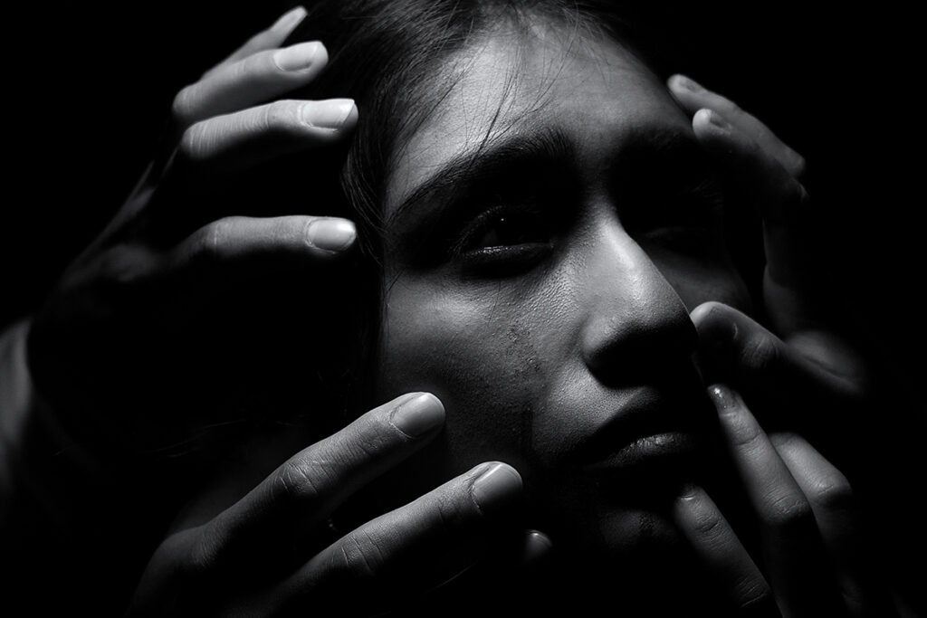 A Black and White image of a Southeast Asian woman looking up, while the hands of many people clutch onto her face as if pulling her back.