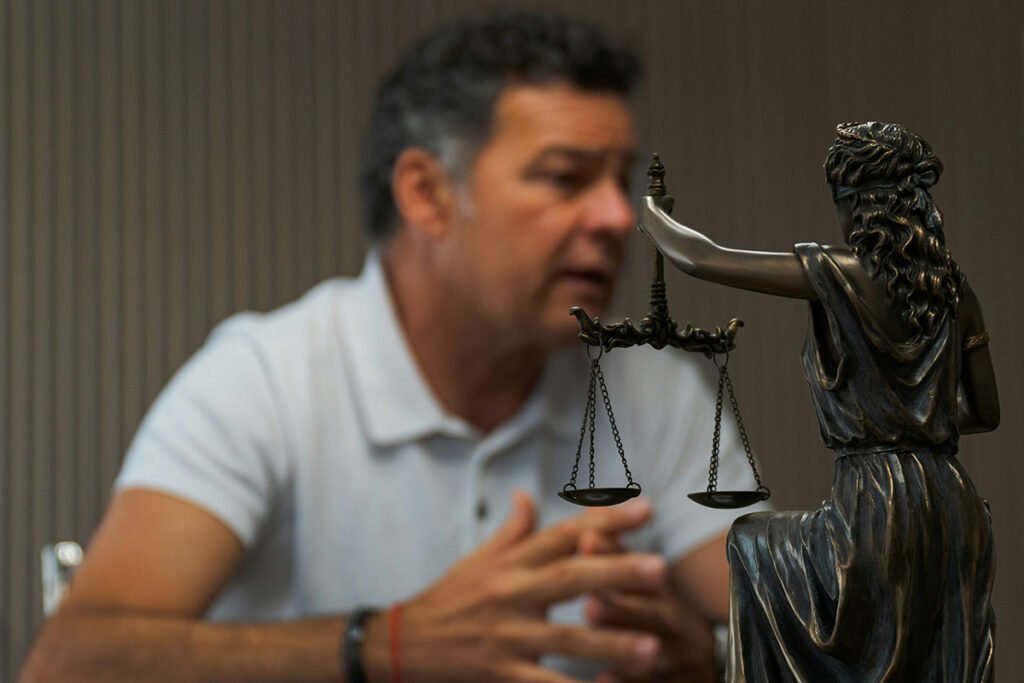 A figurine of lady justice gleams in the foreground, while a man with olive skin and dark hair consults in the background.