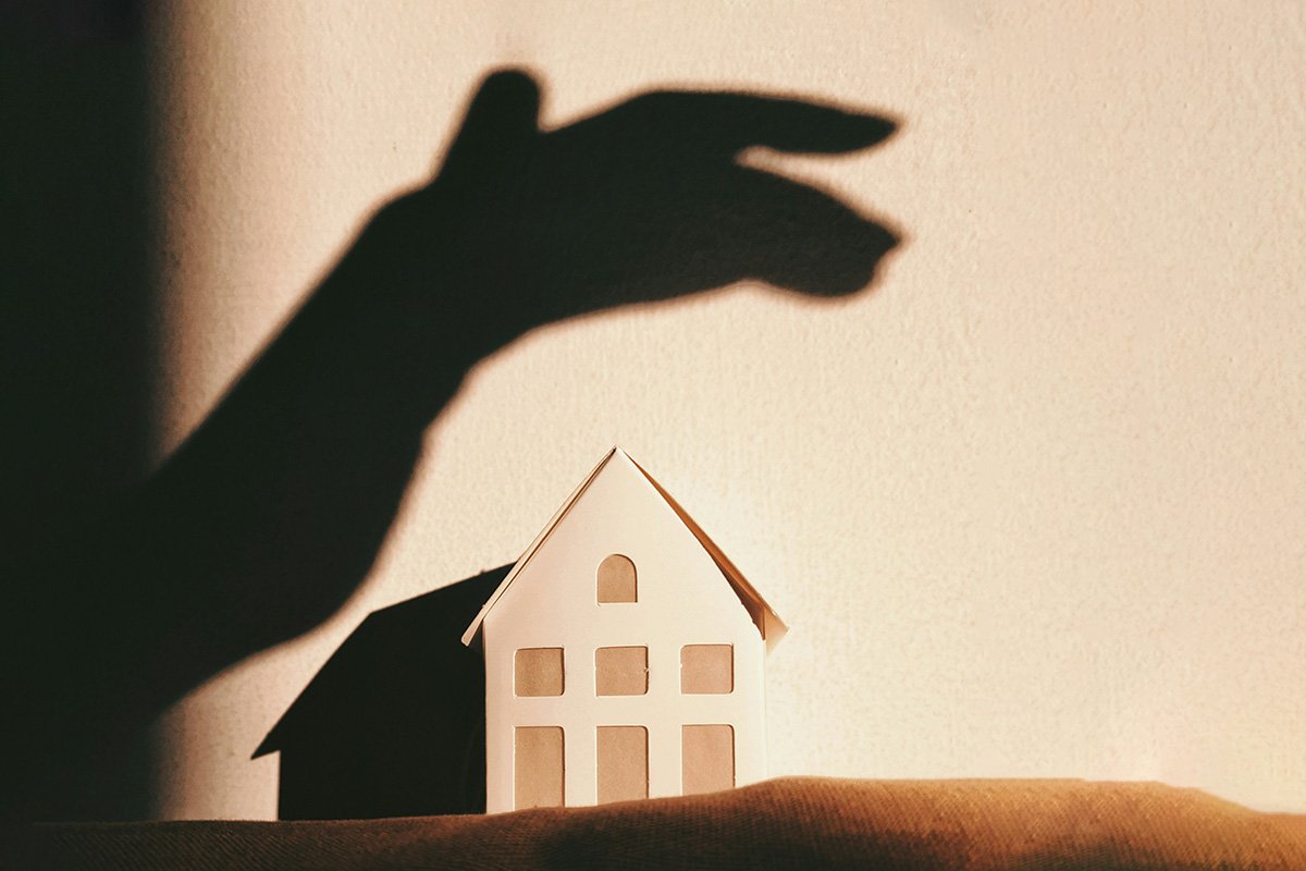 A shadow of a hand looming over a house made of paper, symbolizing the shadow that the Drug War has cast on fair housing.