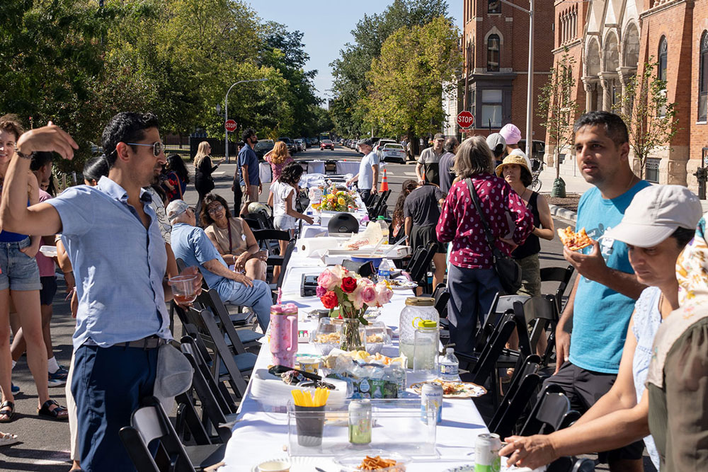 An a group of diverse people sharing an outdoor meal at a long table on a city street. There are flowers, food, and drinks on the table.