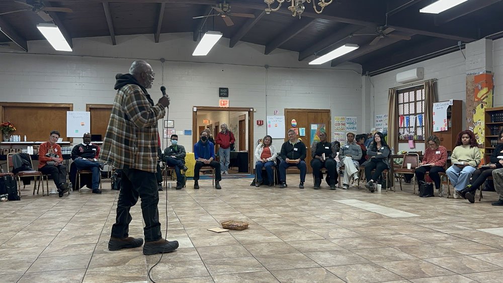 A group of people attending a teach-in, with a Black man holding a mic in the center of the room and people seated around him in a circle.