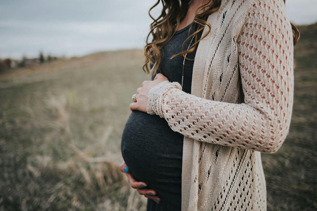 A woman stands in a rural setting with her hands cradling a pregnant belly.