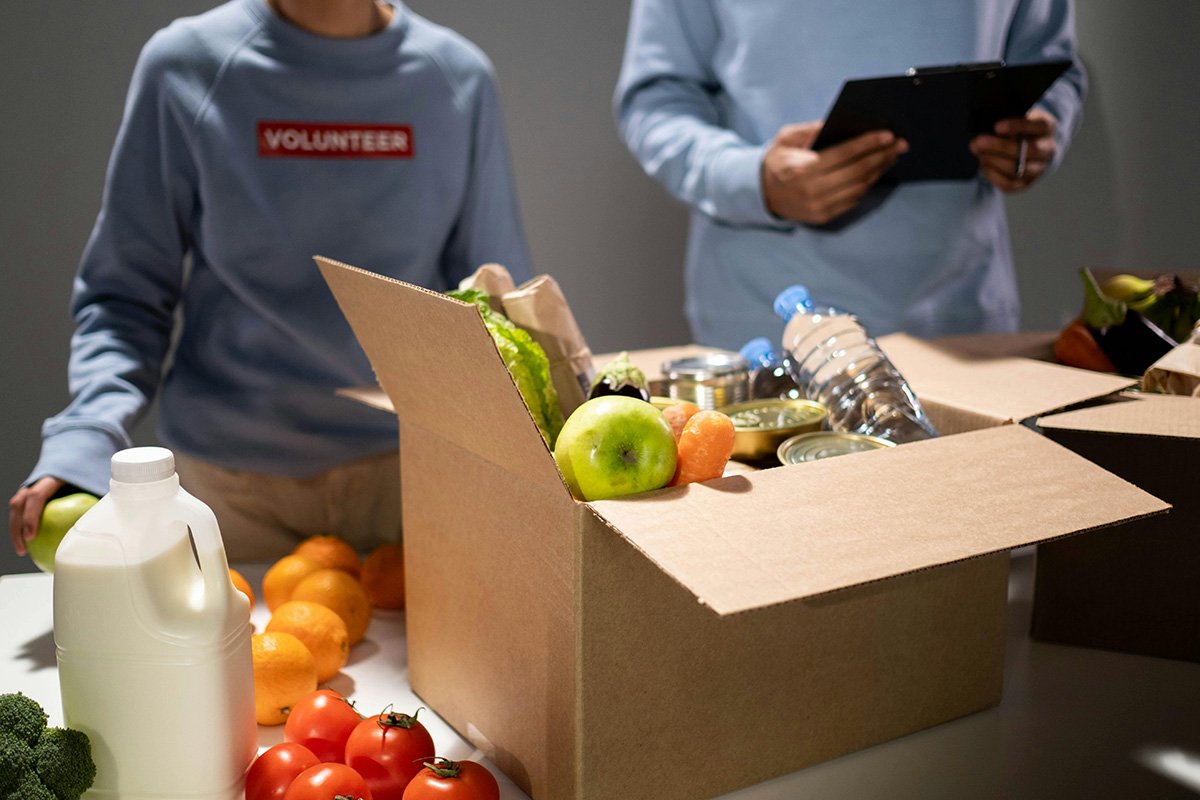 Two people in volunteer shirts standing in front of boxes of produce and donated food, representing the nonprofits who stood in for government responsibility.