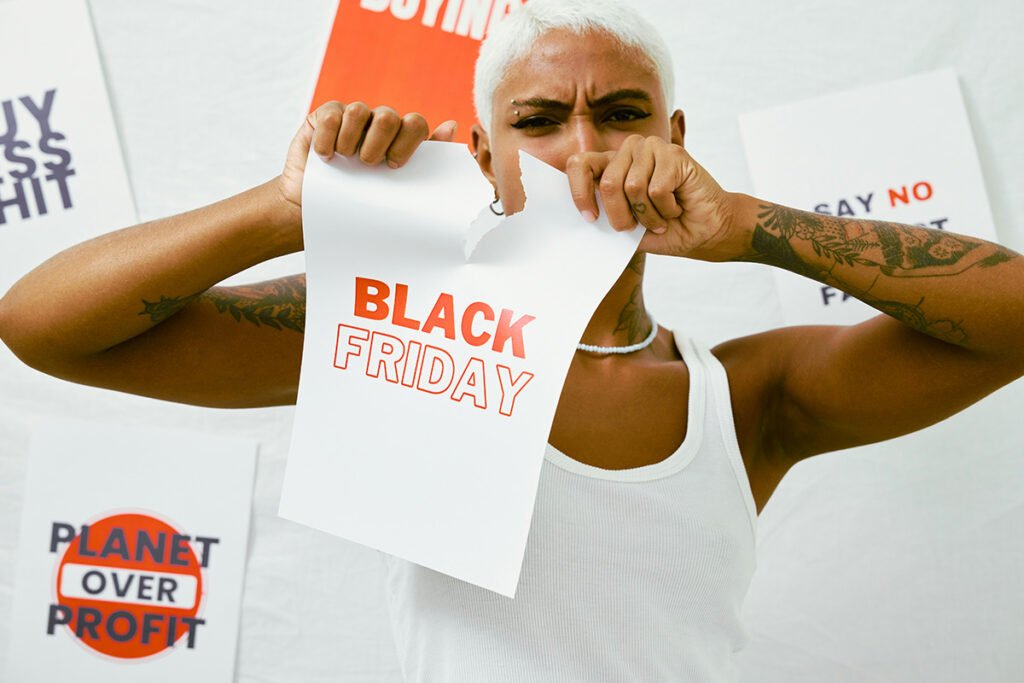 A woman of color with a platinum pixie tearing a paper that says, “Black Friday”. Behind her signs that read, “Buy Less Shit” and “Planet Over Profit”