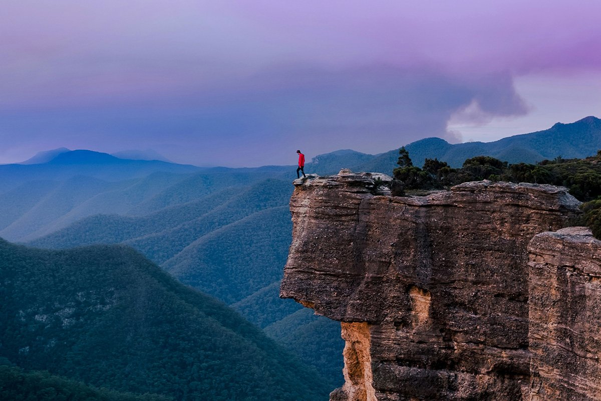 A person looks over the edge of a high cliff in a mountainous landsacpe, symbolizing uncertainty in long-term planning.