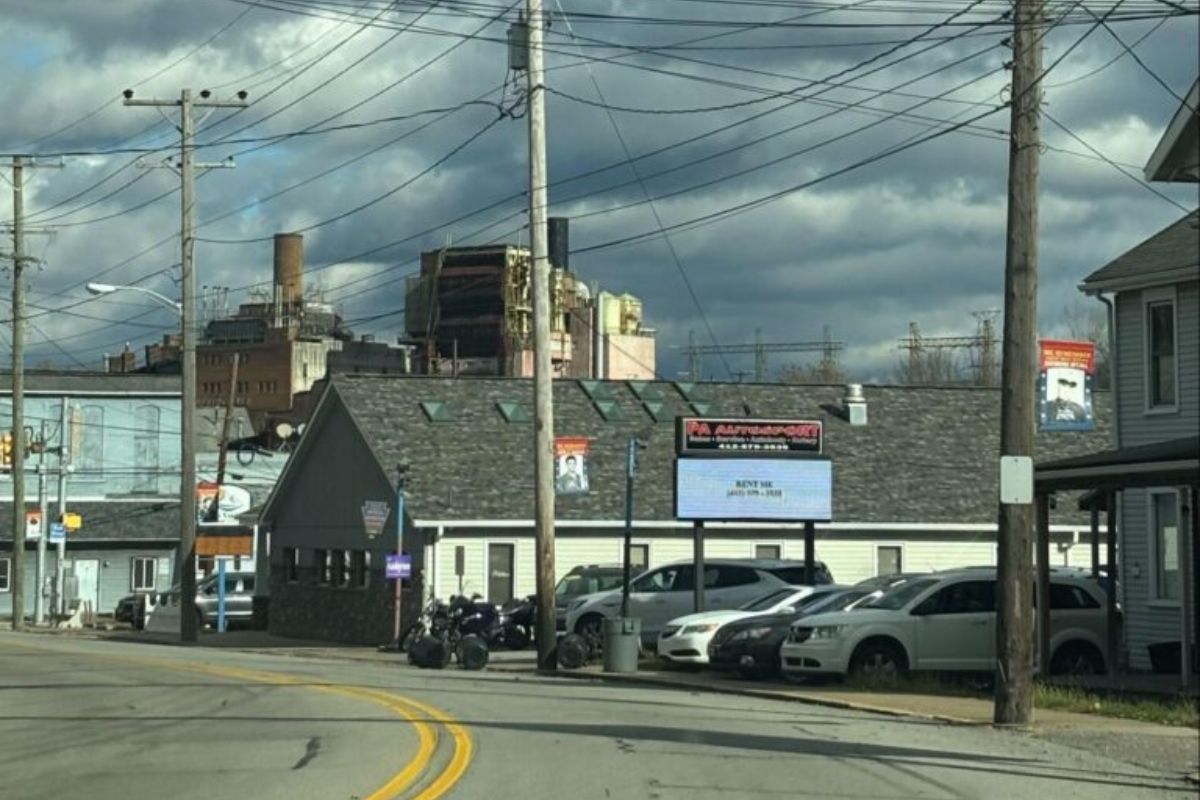 A curving street, buildings, cars, and electrical lines, with a large industrial building in the background.