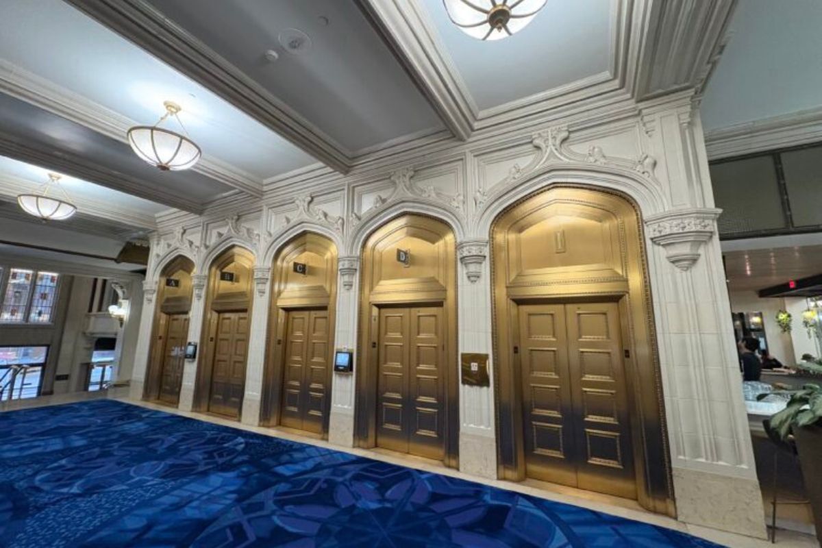 A bank of four golden elevator doors framed by intricate moulding, domed ceiling lights, and deep blue carpeting.