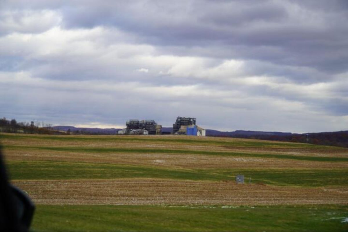 A field of alternating green and brown, with buildings in the distance, under gray cloudy skies.