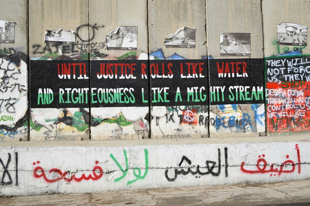 A painted panel of the the Palestine/Israel separation wall., reading “Until justice rolls like water and righteousness like a mighty stream”