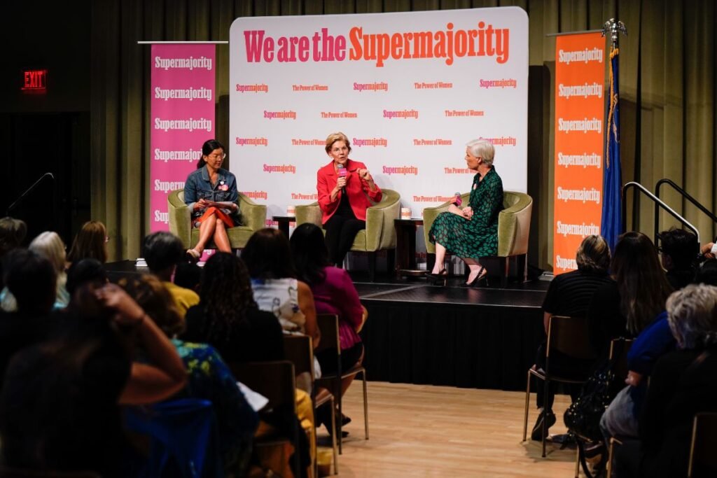 Senator Elizabeth Warren speaks into a microphone in front of a sign reading "We are the Supermajority" while an audience listens.