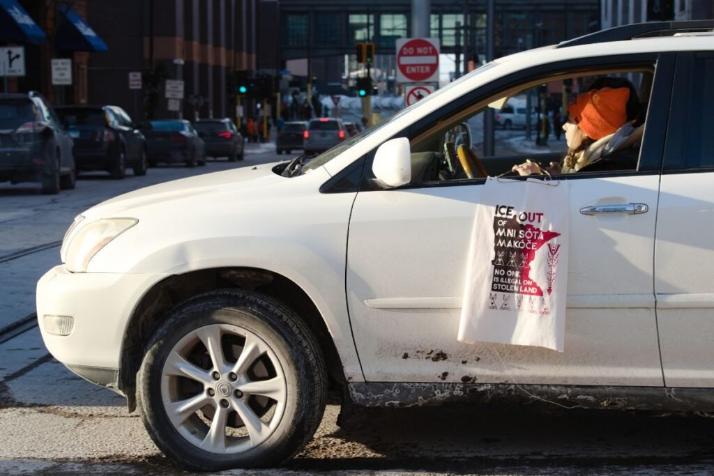 A woman in an orange hat holds a sign out the window of a white car that reads "ICE Out of Mini Sota...No one is illegal on stolen land"