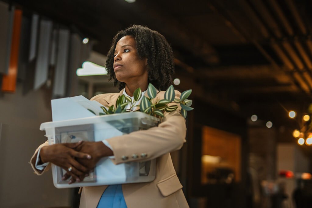 A Black woman in a suit jacket leaving an office with her personal belongings in a plastic bin after a layoff.