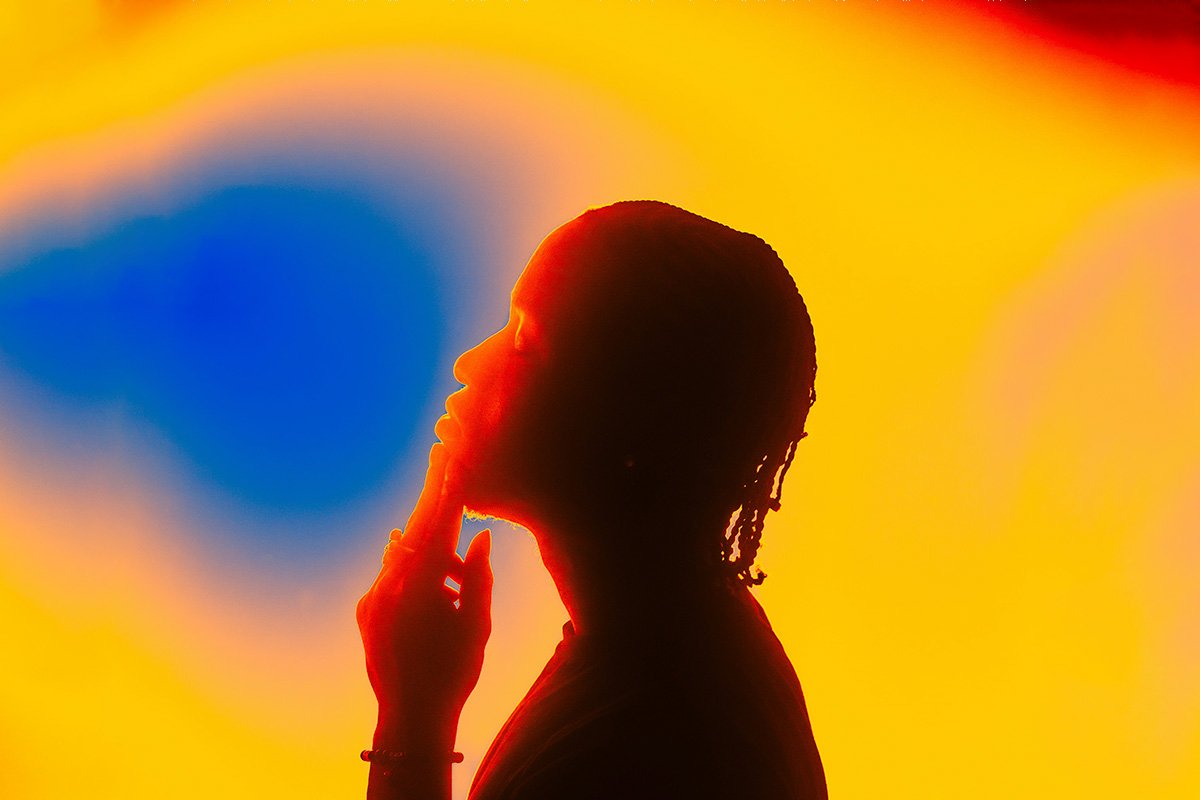 A Black man with cornrows lifts his head and closes his eyes. Behind him, a background glows in a rainbow of color.