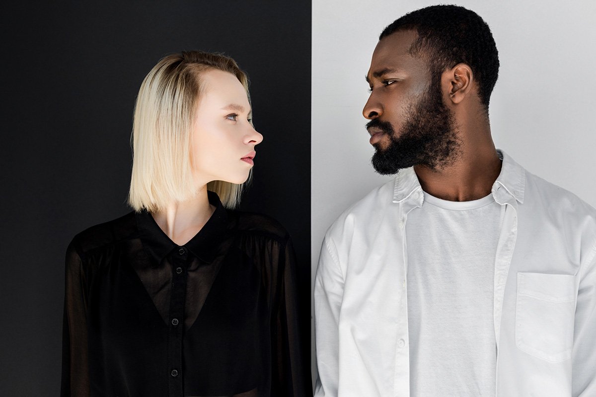 A blonde woman and Black bearded man look each other directly in the eye. The background is split into white and black panels, emphasizing contrast.
