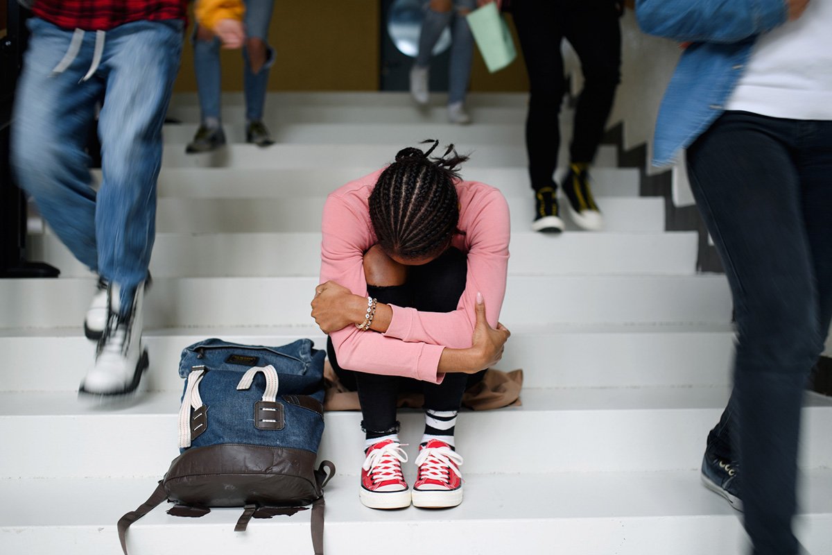 A young Black student with braids sitting on a step with her head tucked in her arms, as other students walk around her.