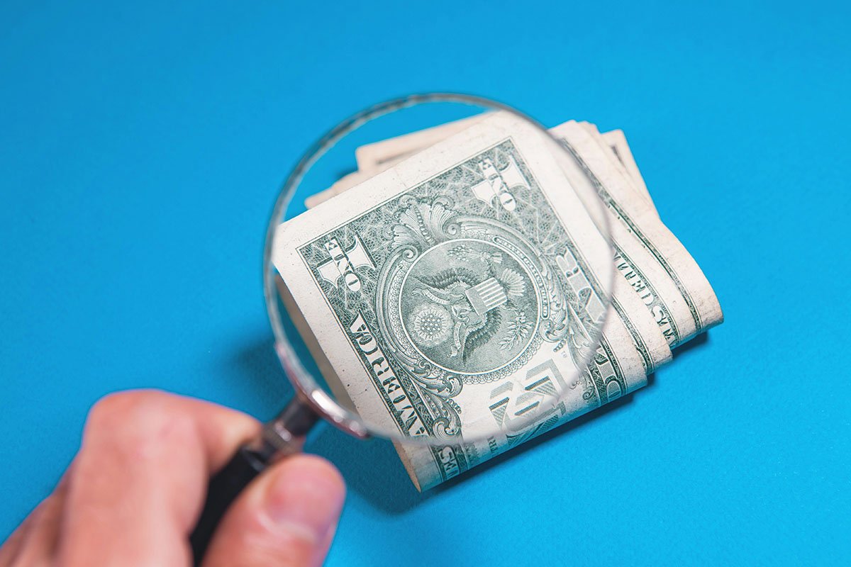 A magnifying glass positioned over a stack of dollar bills, symbolizing the need to take a closer look at public funds.