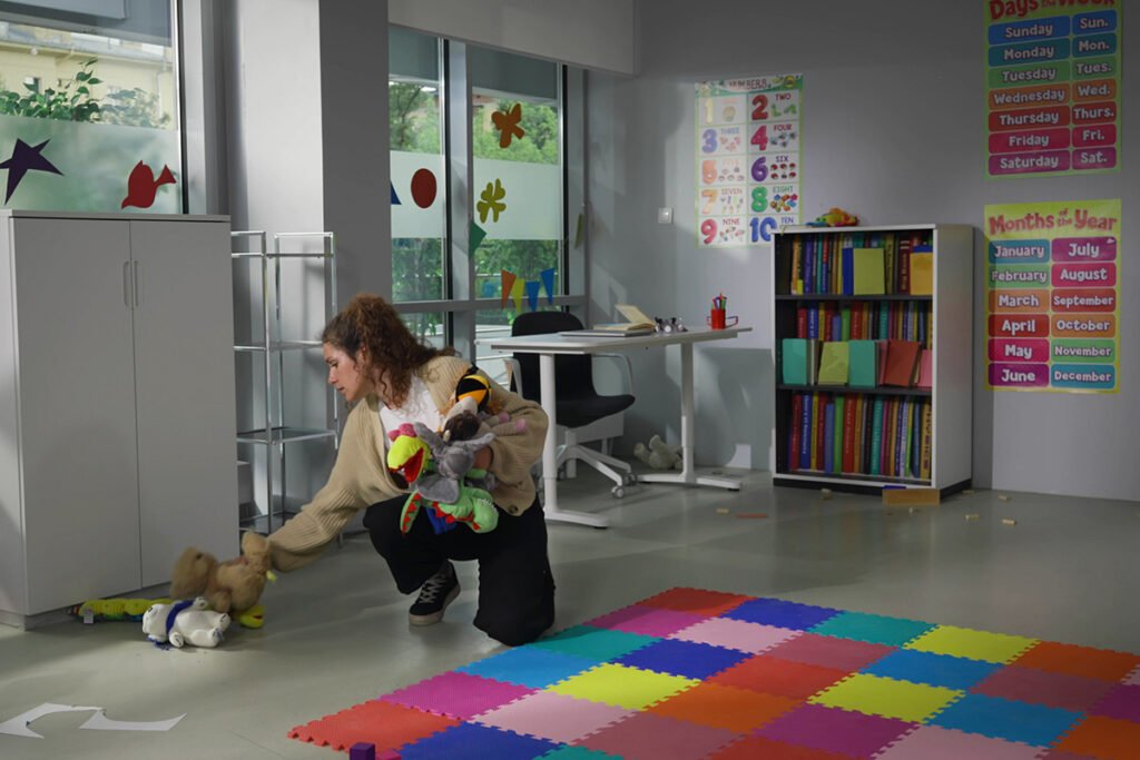 An overwhelmed teacher bending down to pick up toys in an empty elementary classroom.