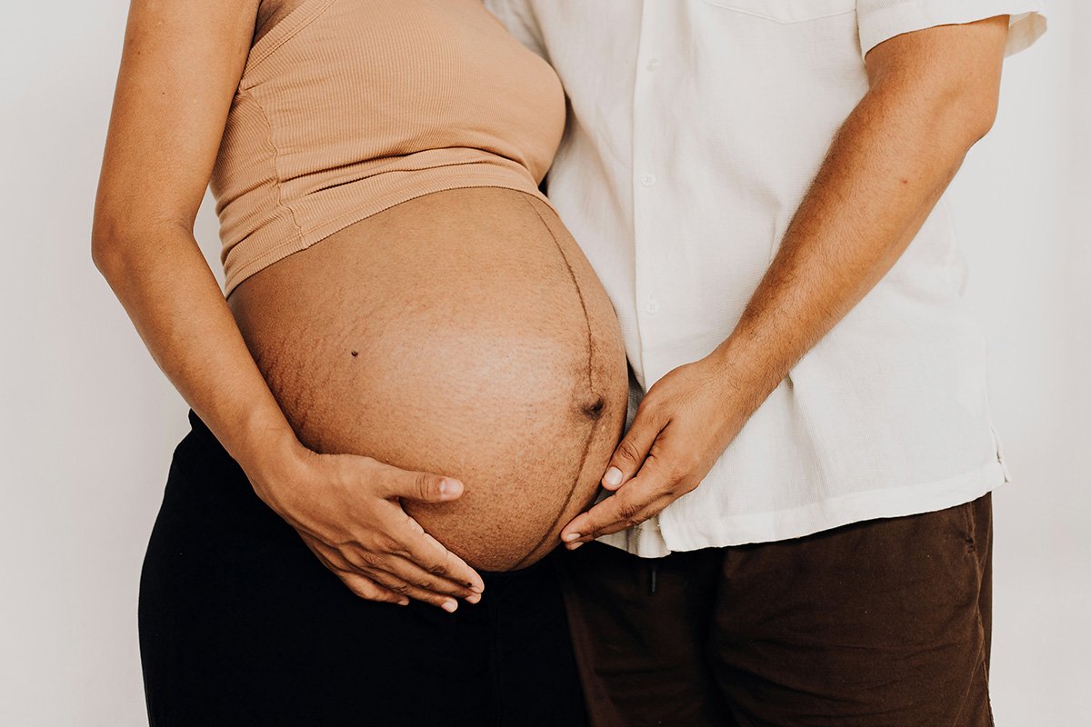 A woman with brown skin and a bare pregnant belly standing with a man who is cradling her belly.