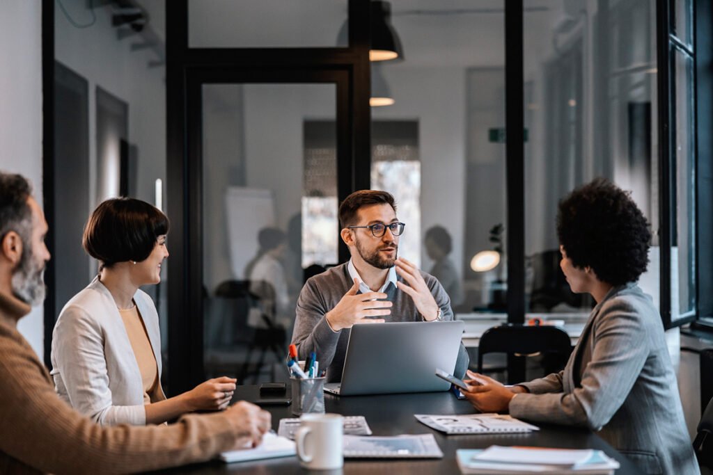 A group of professionals sit around a conference table in an office setting. They listen to a man talk.