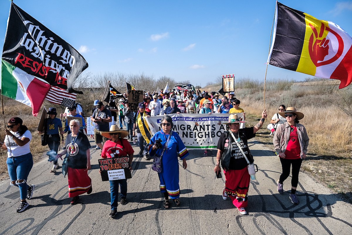 A group of protestors walk down a road together holding flags and signs that read, “Chingo La Migra” and “Immigrants are our neighbors”