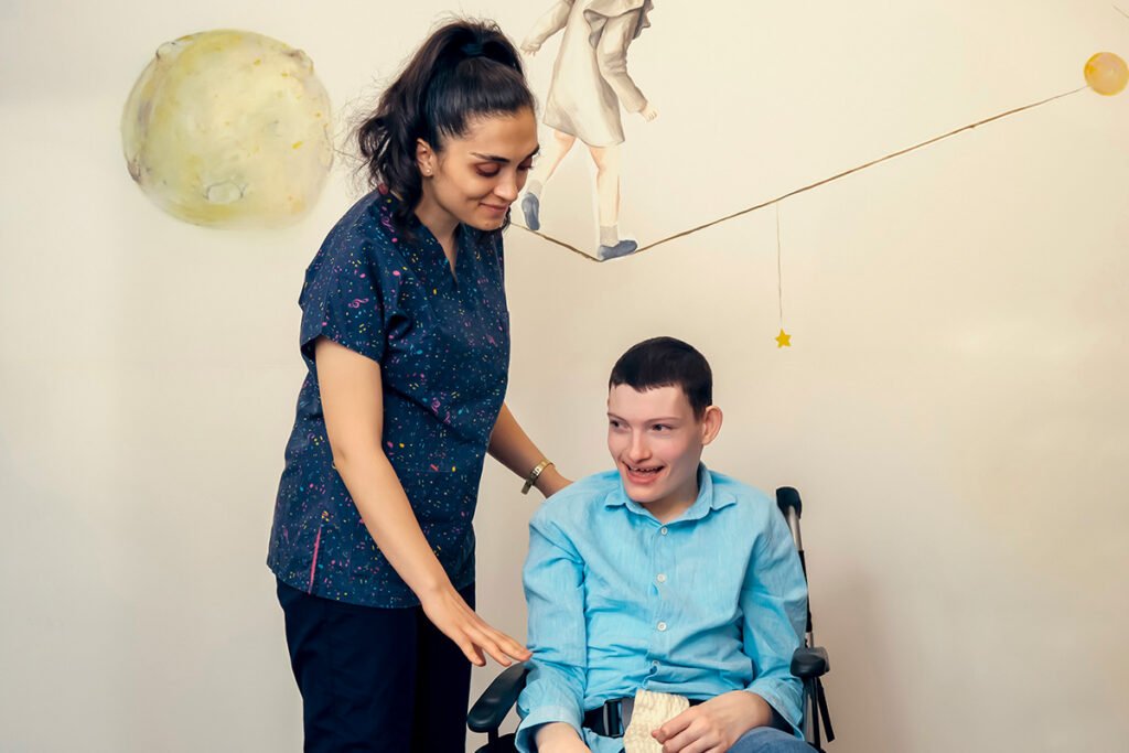 A teenage boy in a wheelchair smiles and looks away, as a caretaker places a hand on his arm. Behind him, there is a mural with a girl walking on a tightrope from the moon to a star.