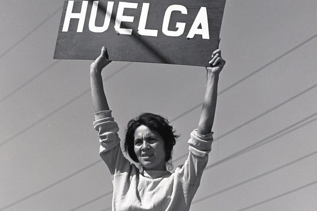 A black and white photo of Dolores Huerta in 1965, holding a sign that reads “HUELGA,” Spanish for “STRIKE,” as she led the farmworkers movement.