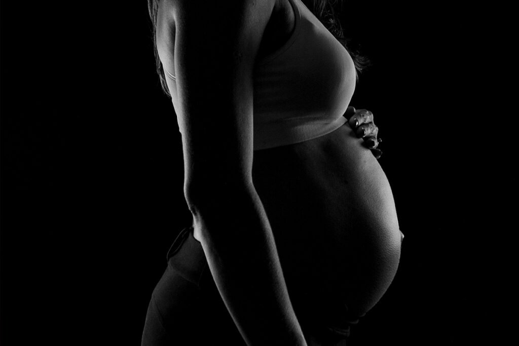 A black and white photo of a woman holding her pregnant belly in low lighting.