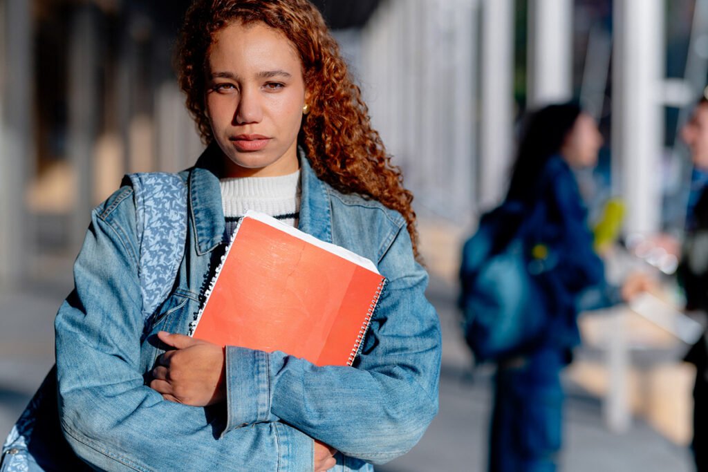 A young Latina student with curly long hair holding a notebook and looking at the camera sullenly.