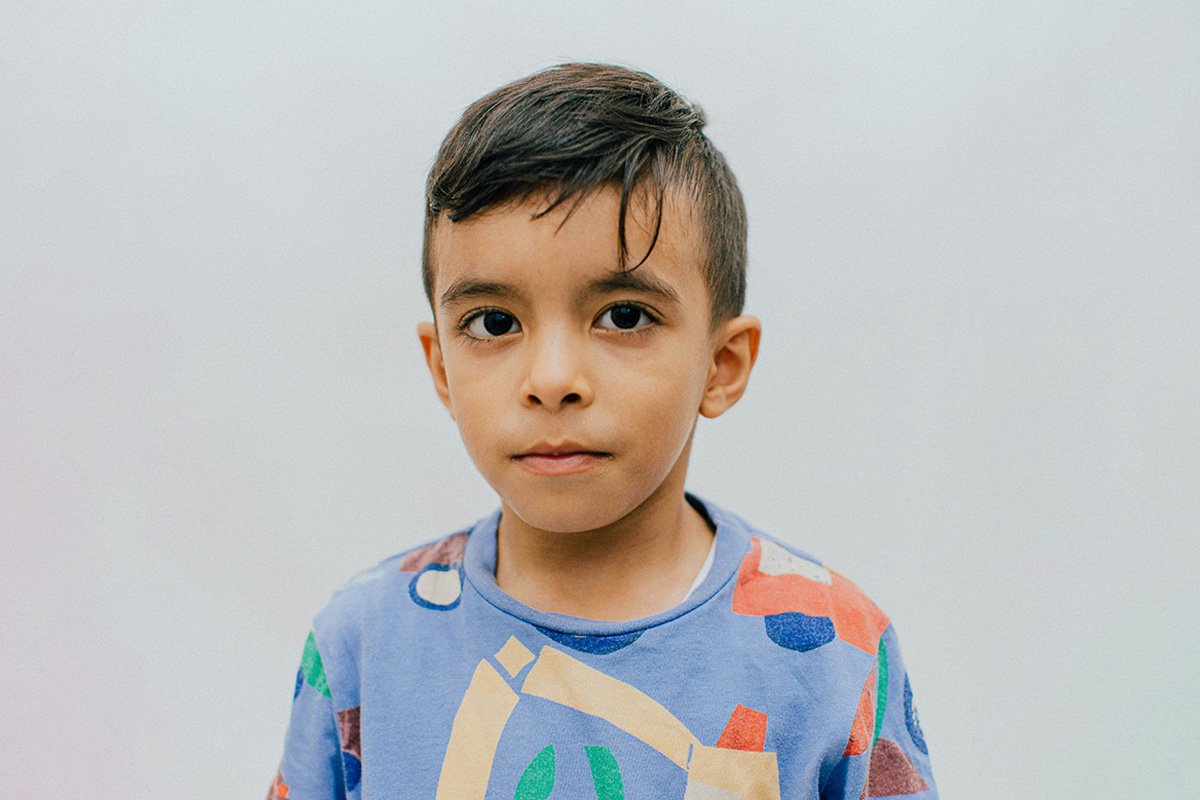A young, Brown child looks into the camera with a reserved expression. He wears a blue shirt with a playful truck print.