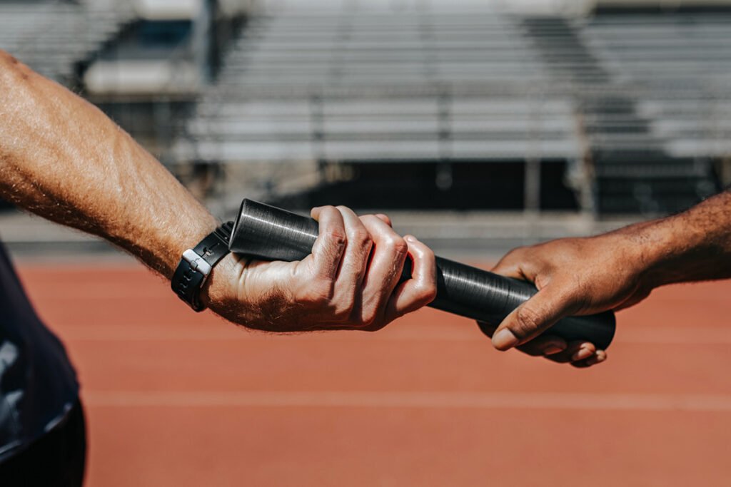 A relay runner passing a baton forward to another runner, representing continuity and responsibility passed forward.