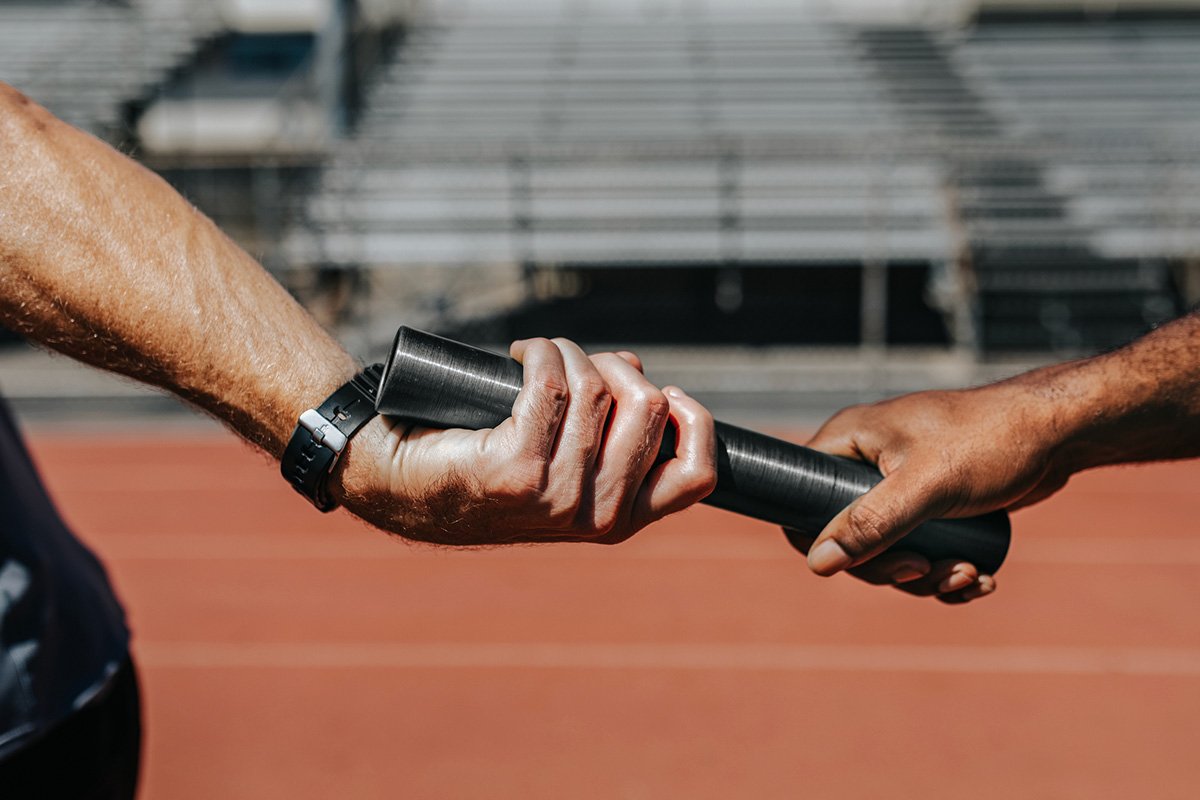 A relay runner passing a baton forward to another runner, representing continuity and responsibility passed forward.