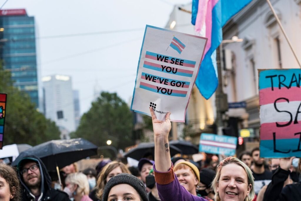 A person at a march holds a sign with the blue, pink, and white trans flag that reads “We See You, We’ve Got You”